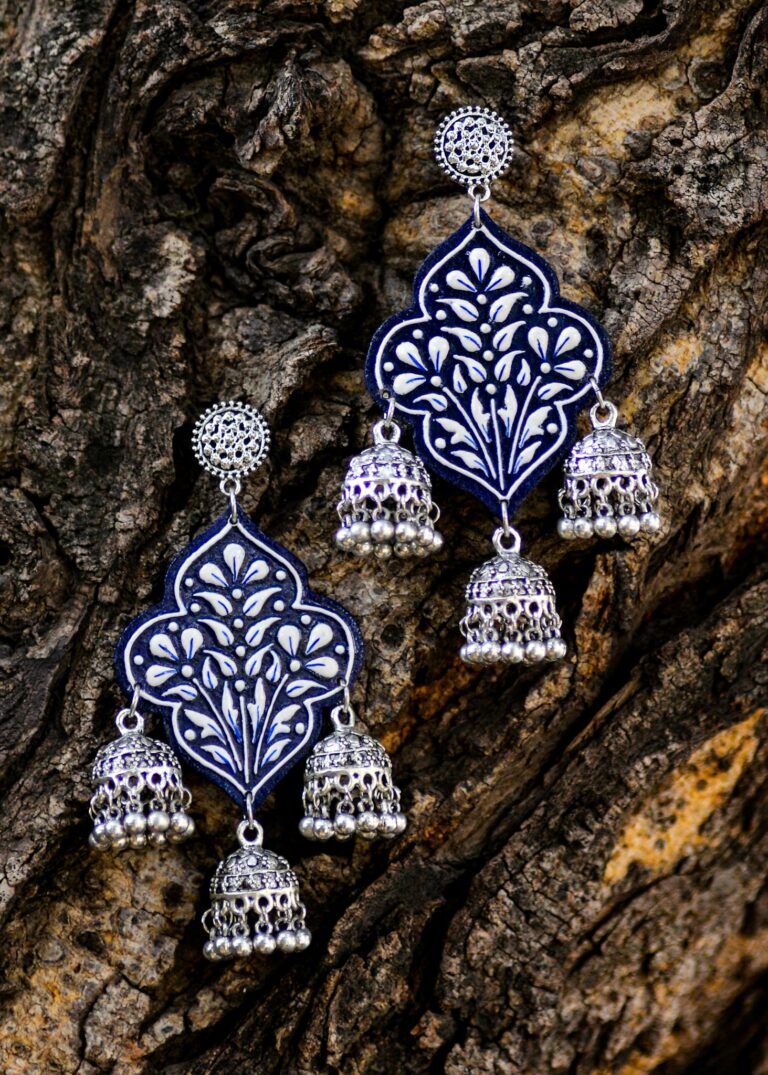 Close-up of ornate silver and blue earrings hanging against textured tree bark, showcasing intricate design.